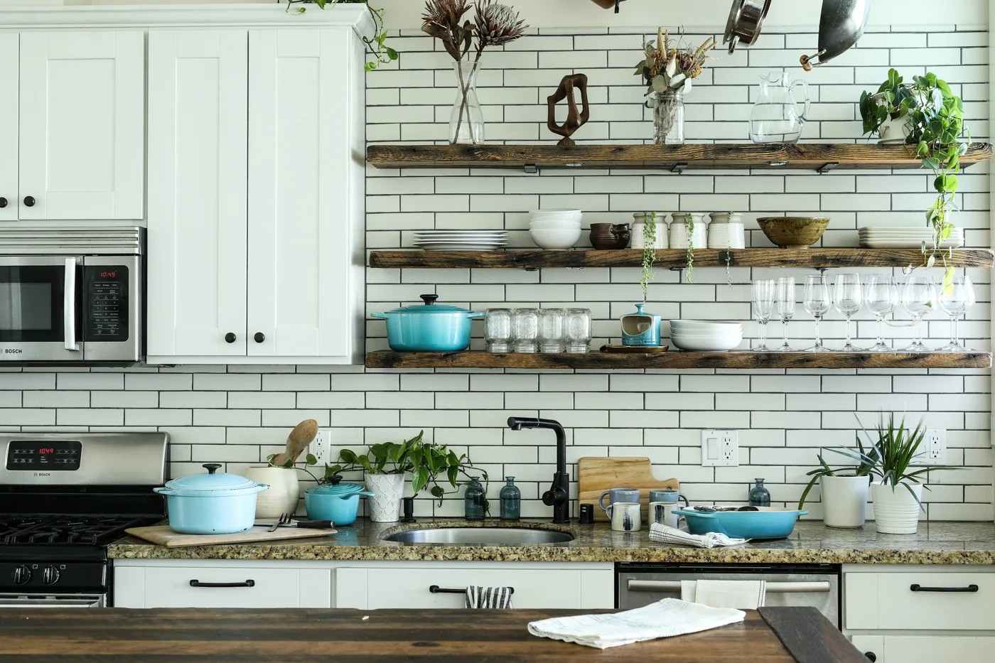 White kitchen cabinets beside open wood shelves, a subway-tile backsplash, and a stone-look counter.