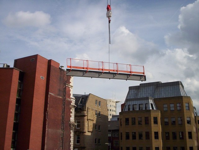 A crane lifts a steel beam with orange edge protection above a construction site among city buildings.